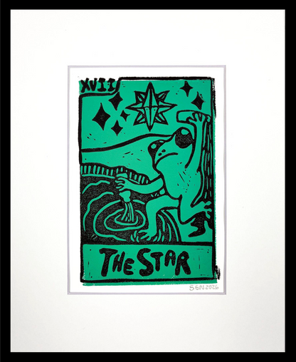 A framed art print featuring a stylized green and black illustration of a frog holding a staff, with the text 'THE STAR' on the left side, set against a white mat and black frame.